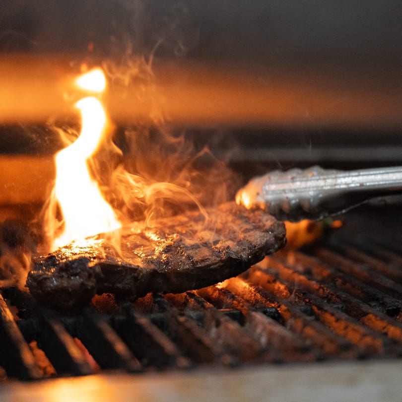 A thick steak sizzles on a fiery grill, held by metal tongs.