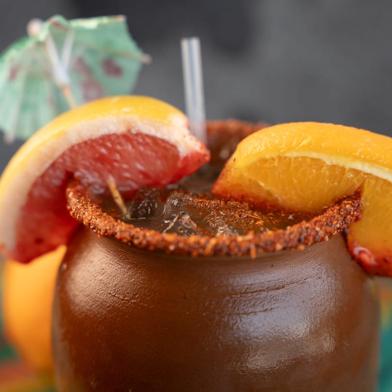 Close-up of a brown clay mug with a dark drink, citrus slices, and a chili-salt rim.
