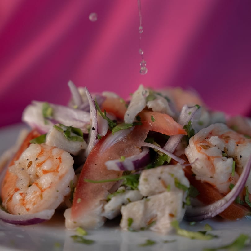 Close-up of ceviche with shrimp, fish, red onion, and tomato, with liquid drizzling from above.