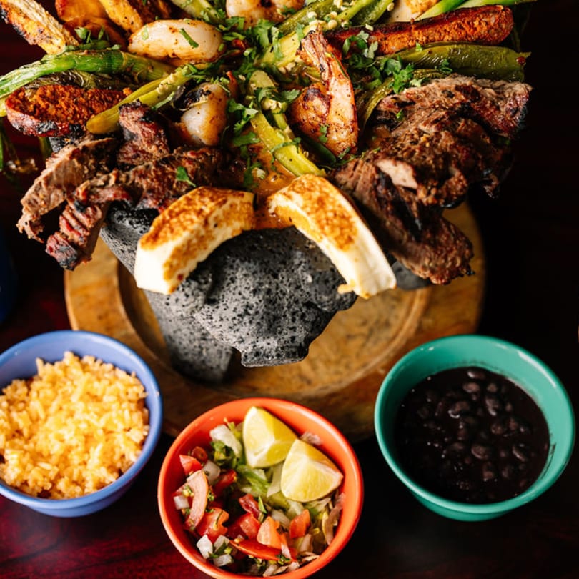 Molcajete dish with grilled steak, shrimp, chorizo, cheese, and vegetables, served with rice, beans, and salsa.