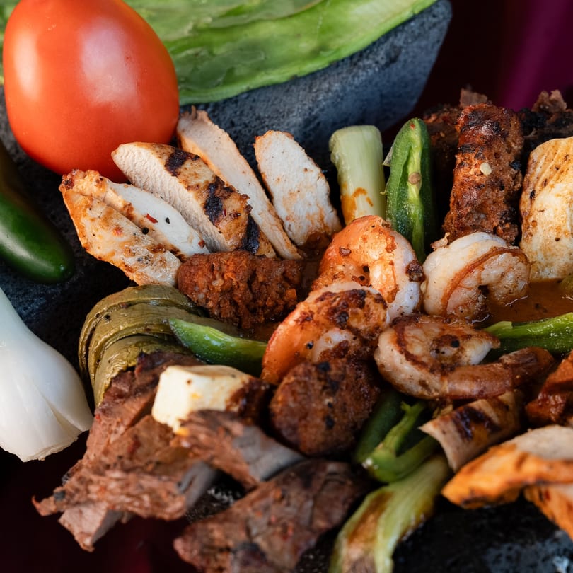Grilled chicken, shrimp, and steak fajitas with vegetables and a tomato on a dark platter.