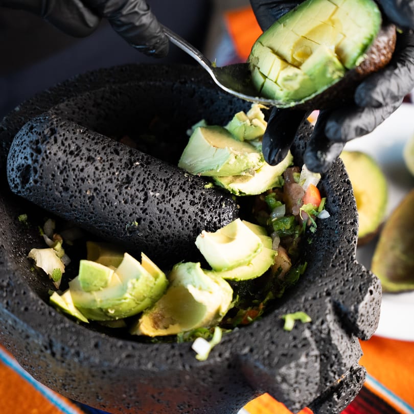 Hands in black gloves spoon diced avocado into a molcajete with guacamole ingredients.