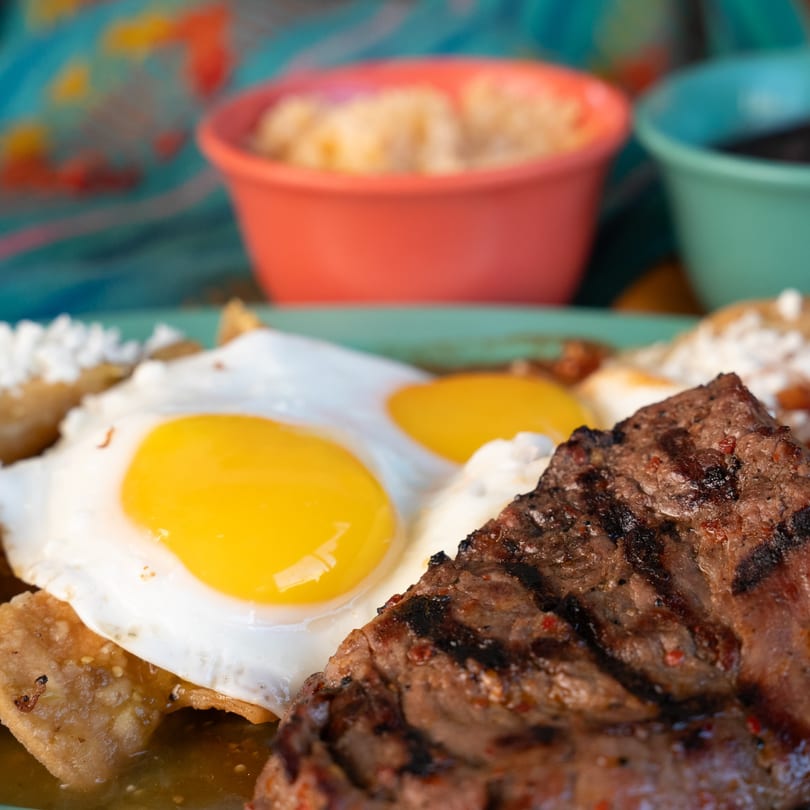Grilled steak and two fried eggs on chilaquiles with side dishes in colorful bowls.
