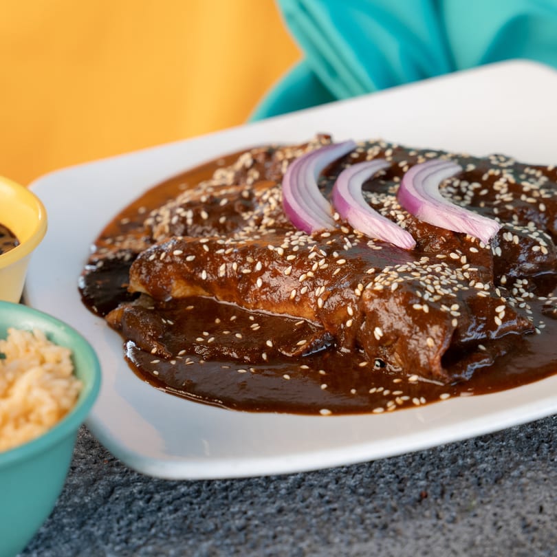 Chicken enchiladas with rich mole sauce, sesame seeds, and red onion slices, served with rice.