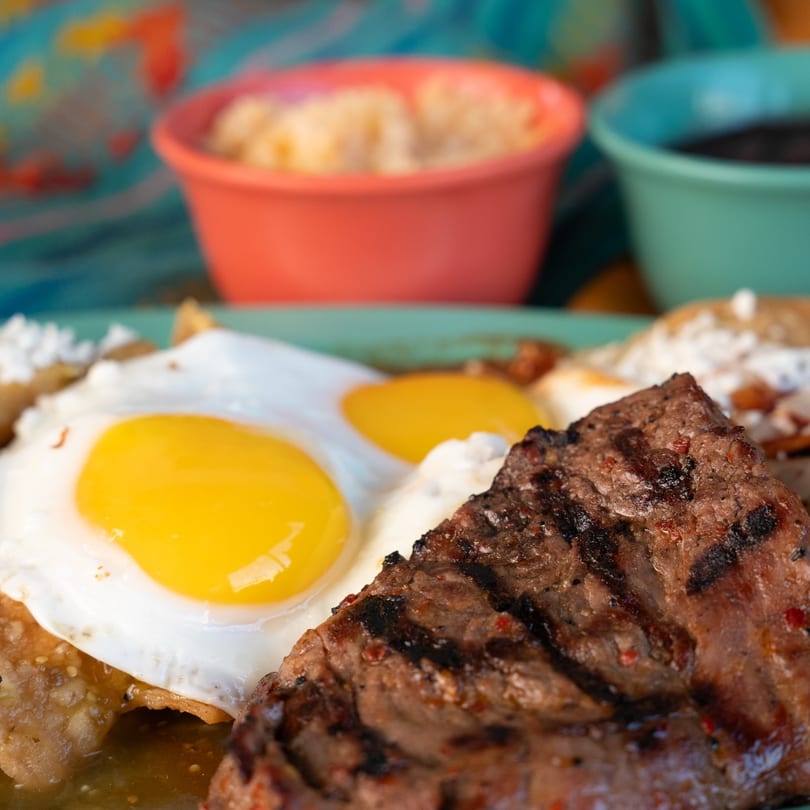 Grilled steak and two fried eggs on a bed of chilaquiles, with side dishes in colorful bowls.