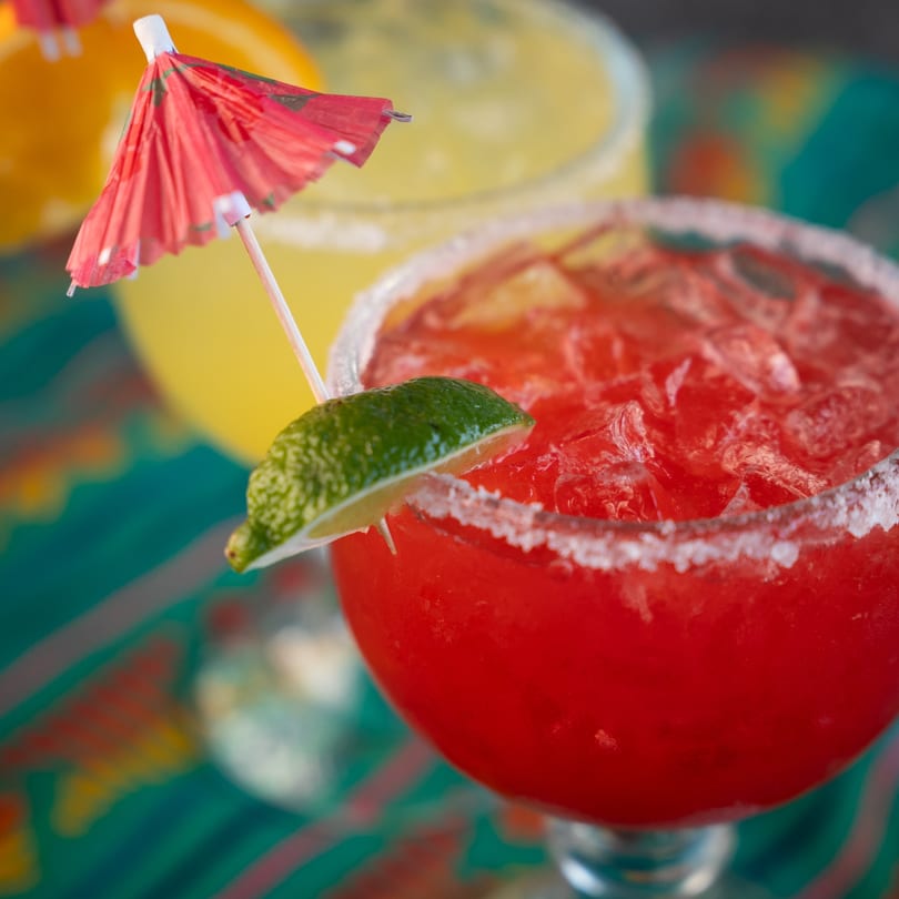 Two margaritas, one red with a lime and umbrella, and one yellow, on a colorful patterned surface.