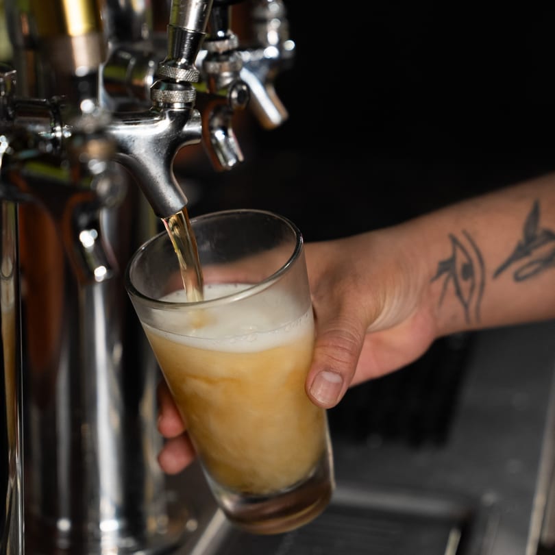 A bartender's tattooed hand holding a glass, filling it with beer from a tap.