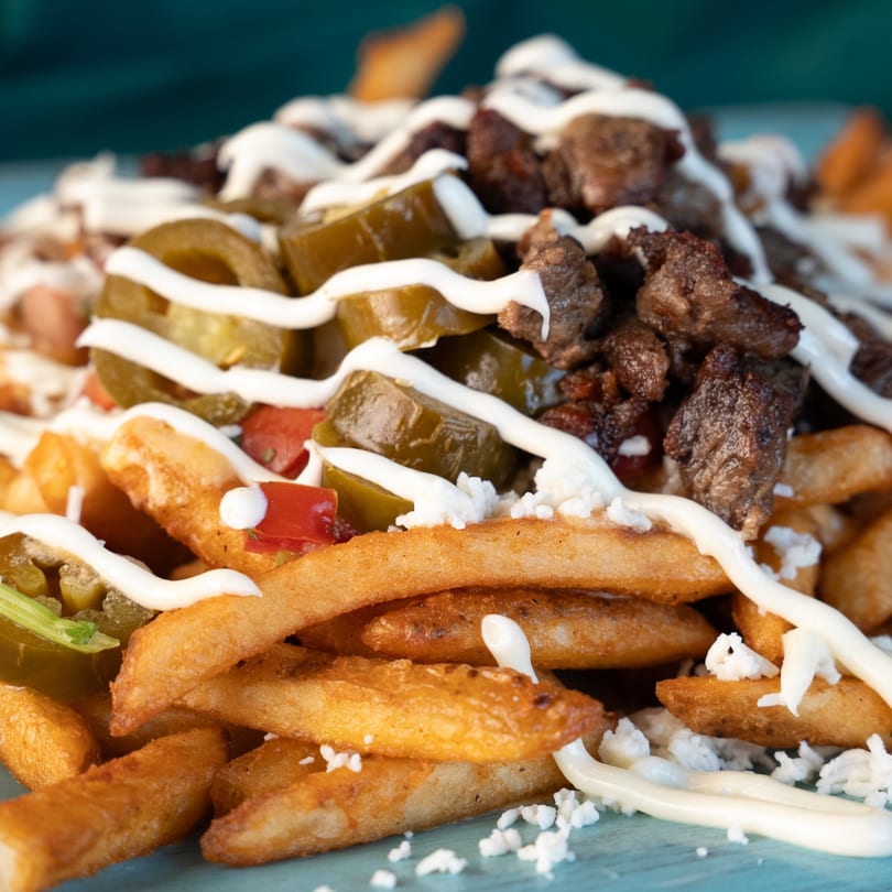 Close-up of golden french fries topped with meat, jalapeños, red peppers, and white sauce.